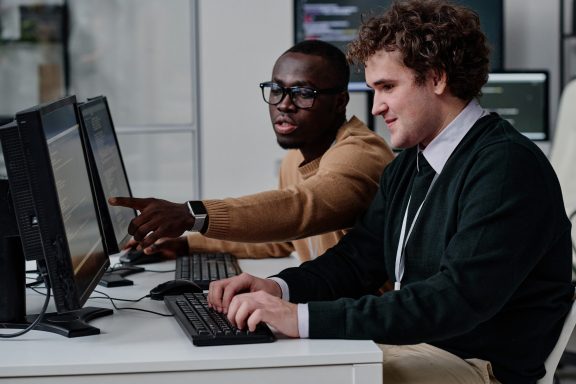 Programmer pointing at computer monitor with codes and talking to his colleague while they working with computer program in team